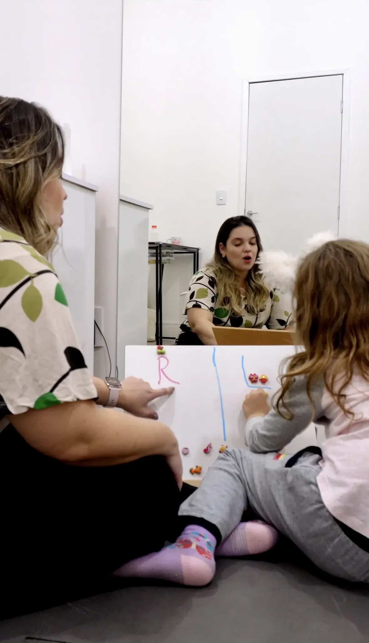 Sessão de fonoaudiologia com a criança e a mãe sentadas no chão, acompanhando a atividade no quadro